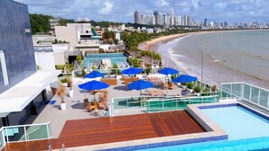 Un área de piscina en la azotea con sillones y sombrillas, con vista a una playa y al horizonte de la ciudad.