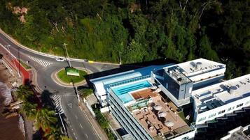 An aerial view of a modern hotel with a rooftop pool, a roundabout, and lush greenery.