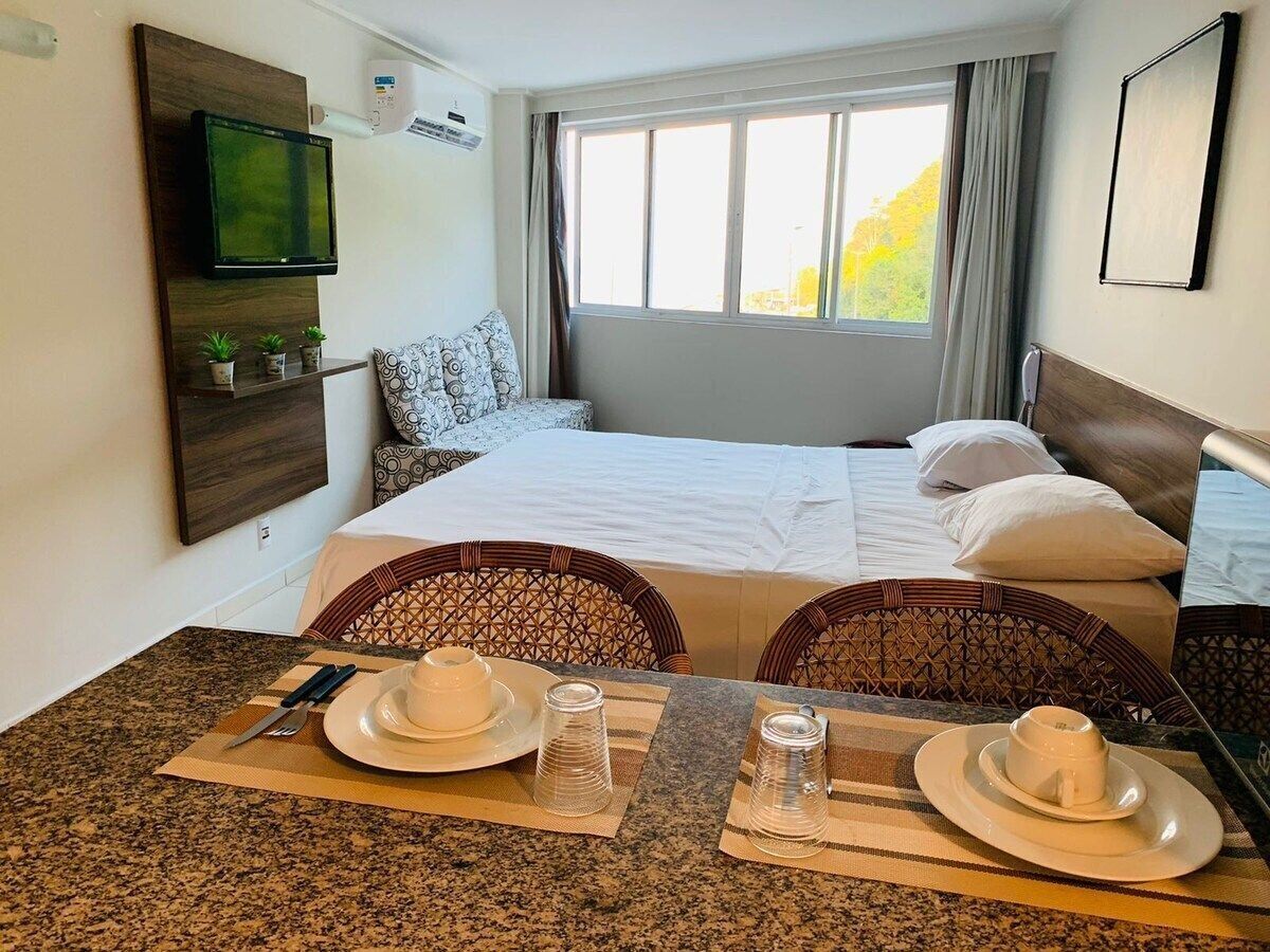 A hotel room with a bed, a TV mounted on the wall, a dining table with plates and cutlery, and a window with curtains.