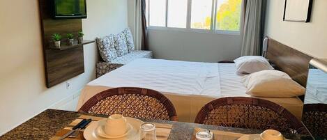 A hotel room with a bed, a TV mounted on the wall, a dining table with plates and cutlery, and a window with curtains.