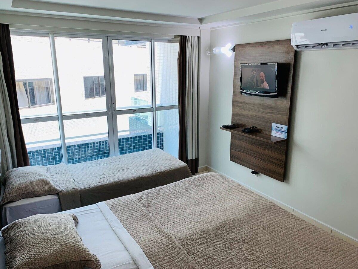Habitación de hotel con dos camas, un televisor montado en la pared y vistas a la ciudad a través de amplias ventanas.