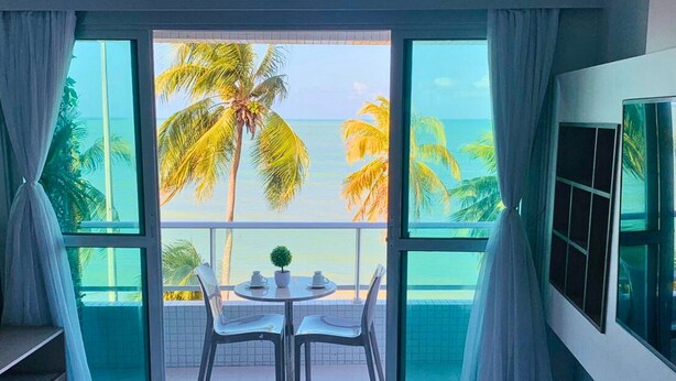 Habitación con un ventanal grande que da a una playa con palmeras y el océano. Hay una cama con cubierta roja, una mesita con sillas y una planta en maceta.