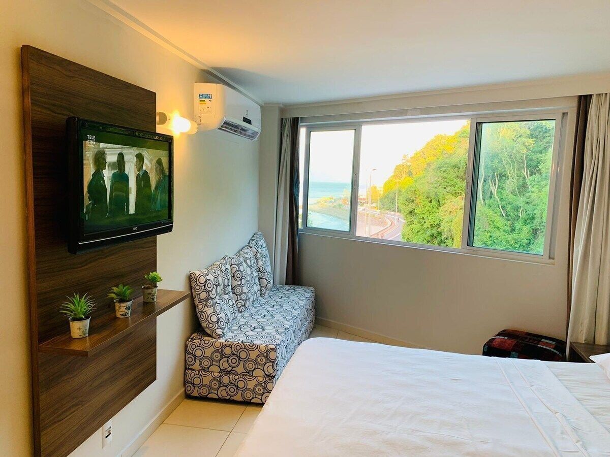 A hotel room with a large bed, a TV mounted on the wall, a window with a view of trees, and a small seating area with cushions.