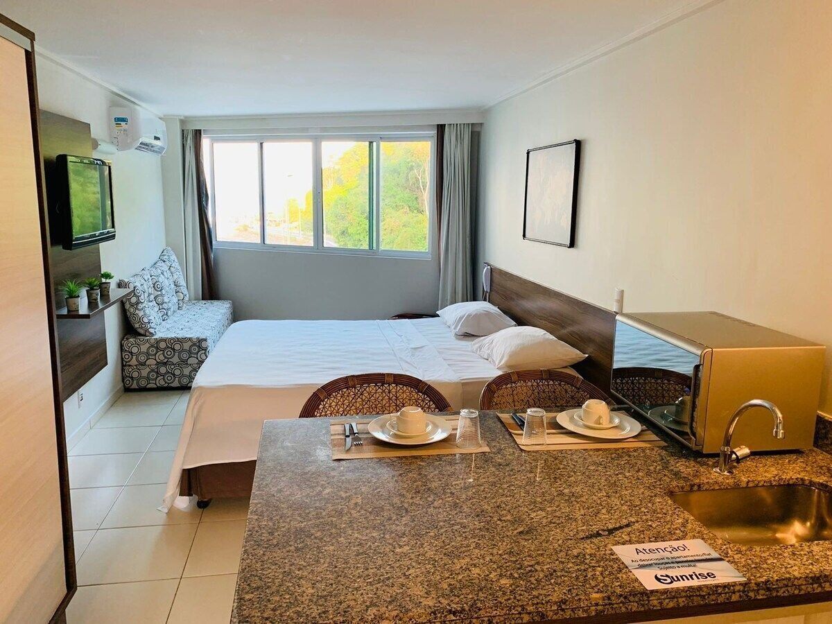 A hotel room with a bed, a small seating area, a kitchenette, and a window with a view of trees.