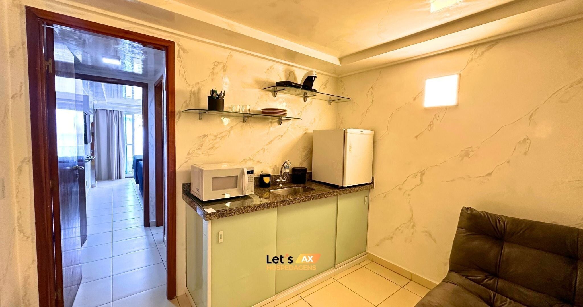 Eine kompakte Küche mit Mikrowelle, Kaffeemaschine und Kühlschrank. Ein Glaskasten über der Arbeitsplatte beherbergt einige Gegenstände. Der Boden ist mit Fliesen ausgelegt und eine Tür führt zu einem Flur.