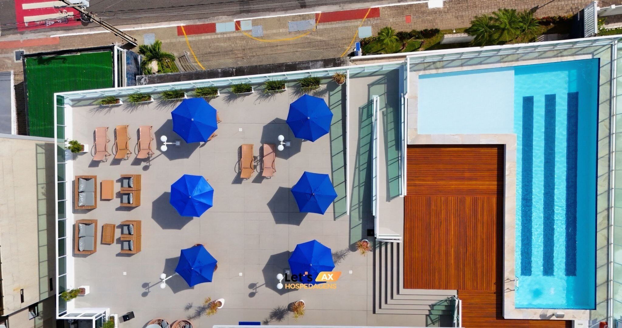 Vista aèria d'una zona de piscina al terrat, amb diverses ombrelles blaves, seients de relax i una porta de fusta.