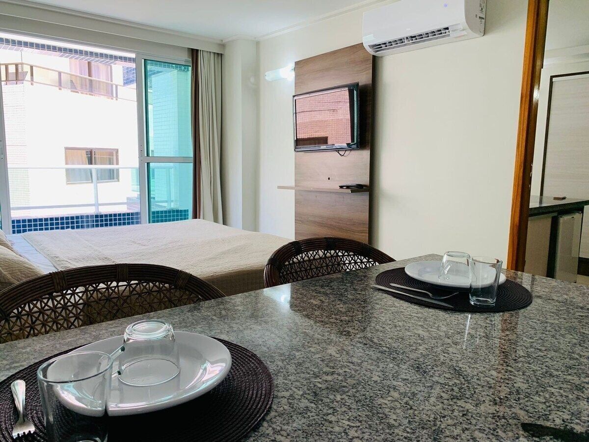 A hotel room with a dining table set for two, a television mounted on the wall, and an air conditioning unit.