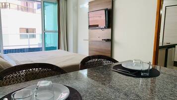 A hotel room with a dining table set for two, a television mounted on the wall, and an air conditioning unit.