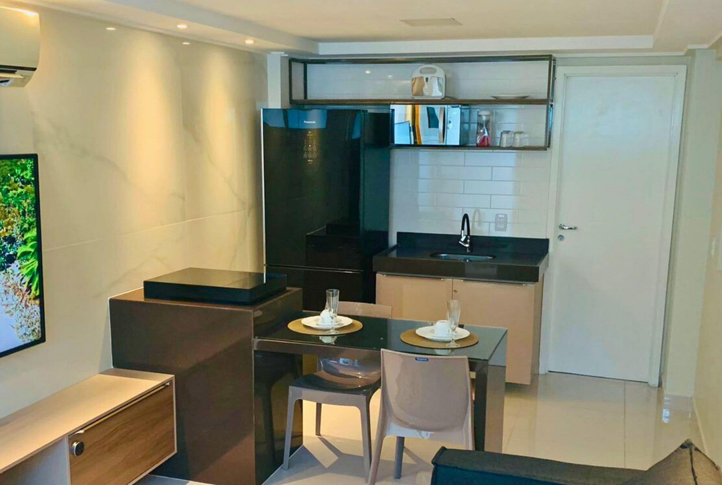 Een kleine keuken met een koelkast, spoelbak en aanrecht. Er is een eettafel met stoelen en een klein kastje.