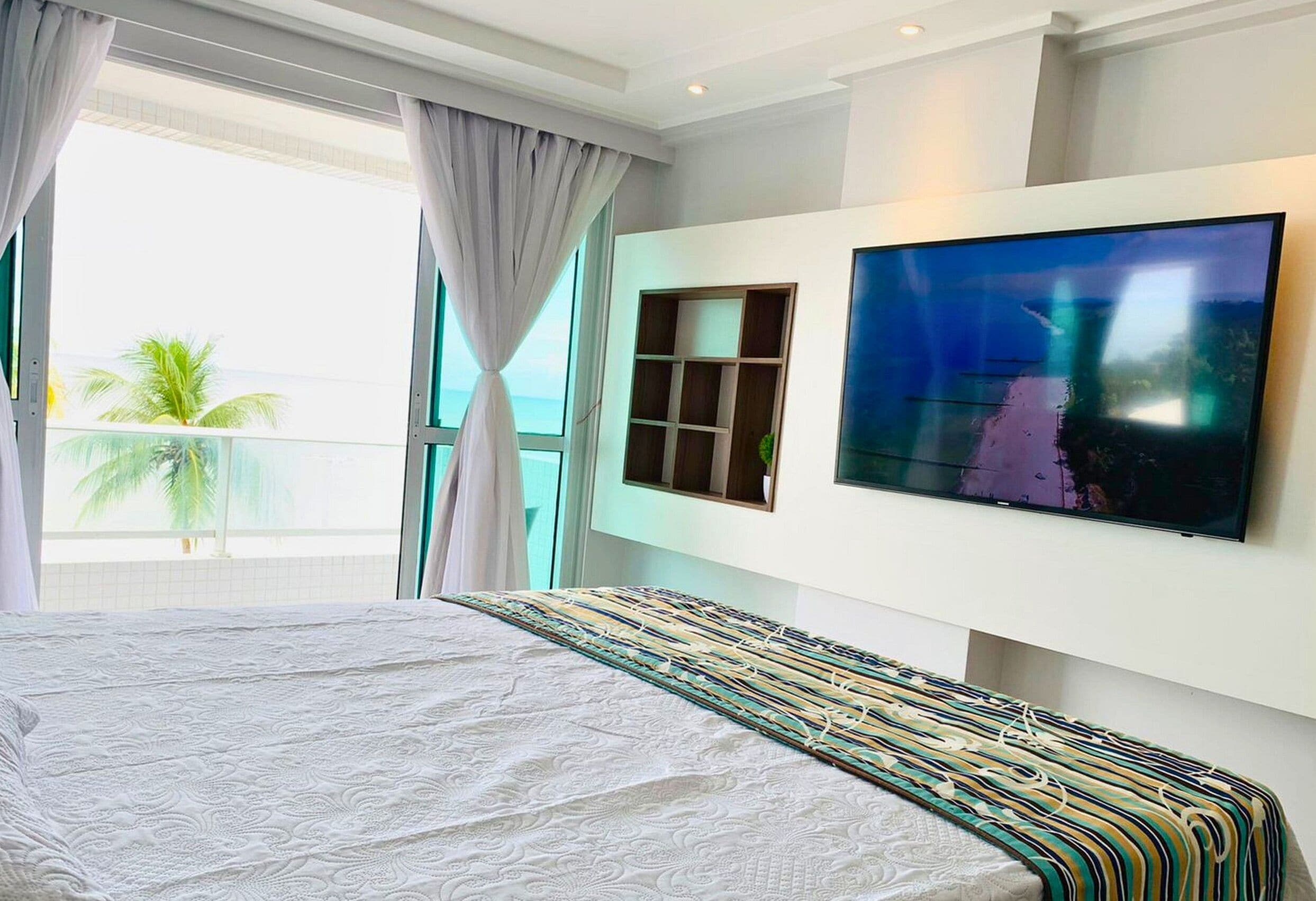 Una camera d'albergo moderna con un letto grande, una TV a parete e un balcone con vista su palme.