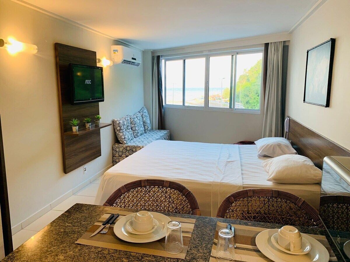 Habitación de hotel con dos camas, un comedor con mesa para dos personas, un televisor montado en la pared y una ventana con vistas a zonas verdes.