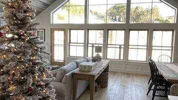 A cozy room with a large window, a wooden ceiling, a Christmas tree, a sofa, a table, and chairs.