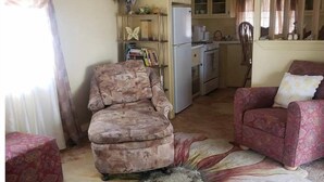 Un saló amb un penjall floral, dues butaques, un sofà, una zona de cuina amb armaris i un frigorífic, i una taula de menjador amb cadires.