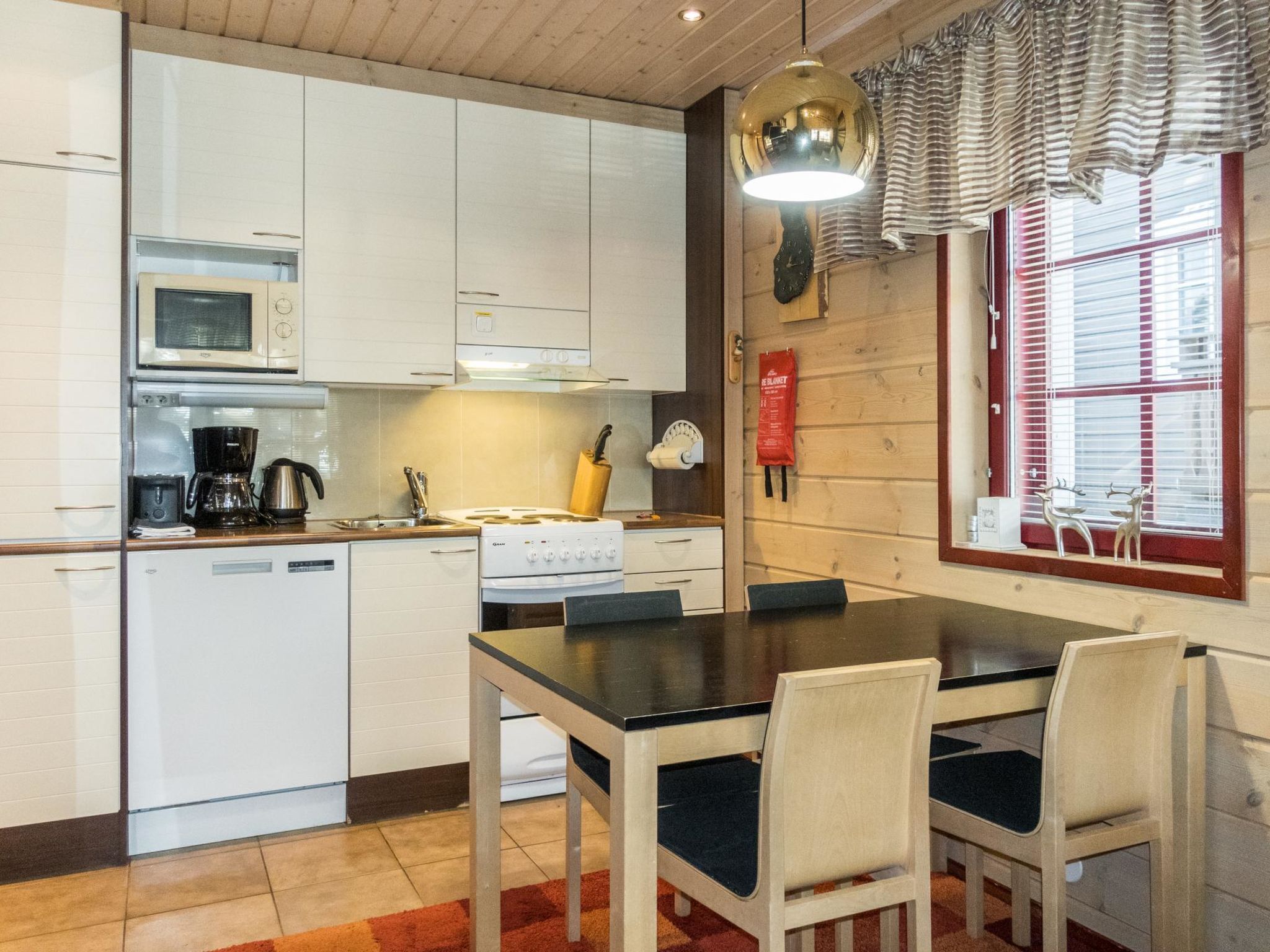 A compact kitchen with white cabinets, a stove, a microwave, and a coffee maker. There is a dining table with chairs and a window with blinds.