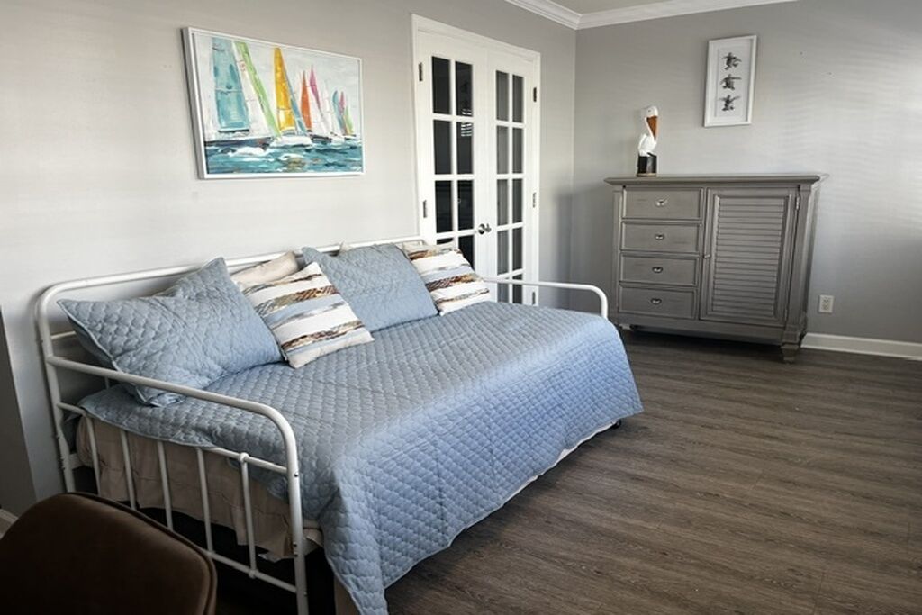 A bedroom with a metal bed frame, a gray quilted bedspread, a white door, a gray dresser, and a painting of sailboats.
