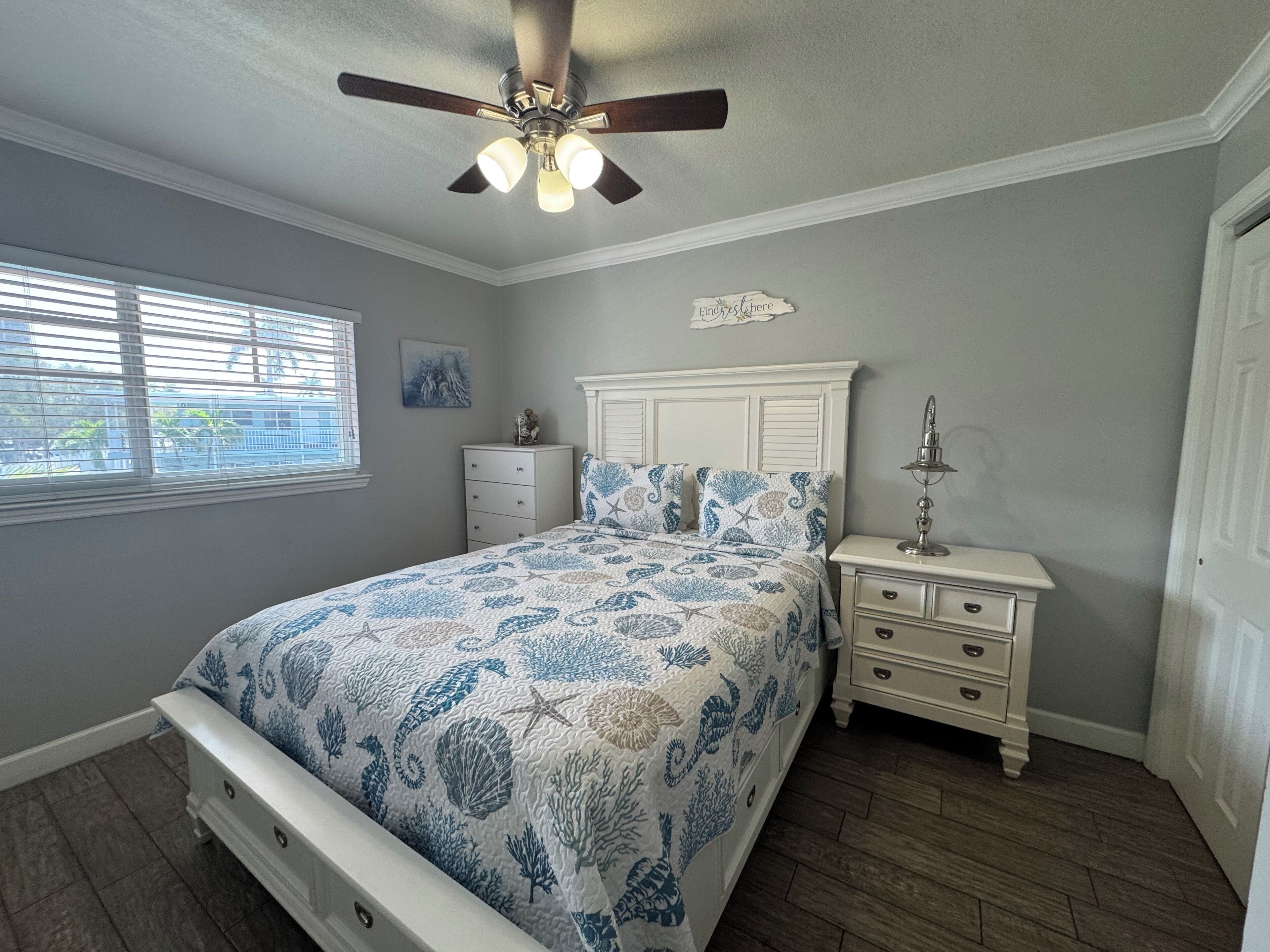 Une chambre à coucher comprenant un lit, une table de chevet, un ventilateur de plafond, une fenêtre avec des stores et une porte.