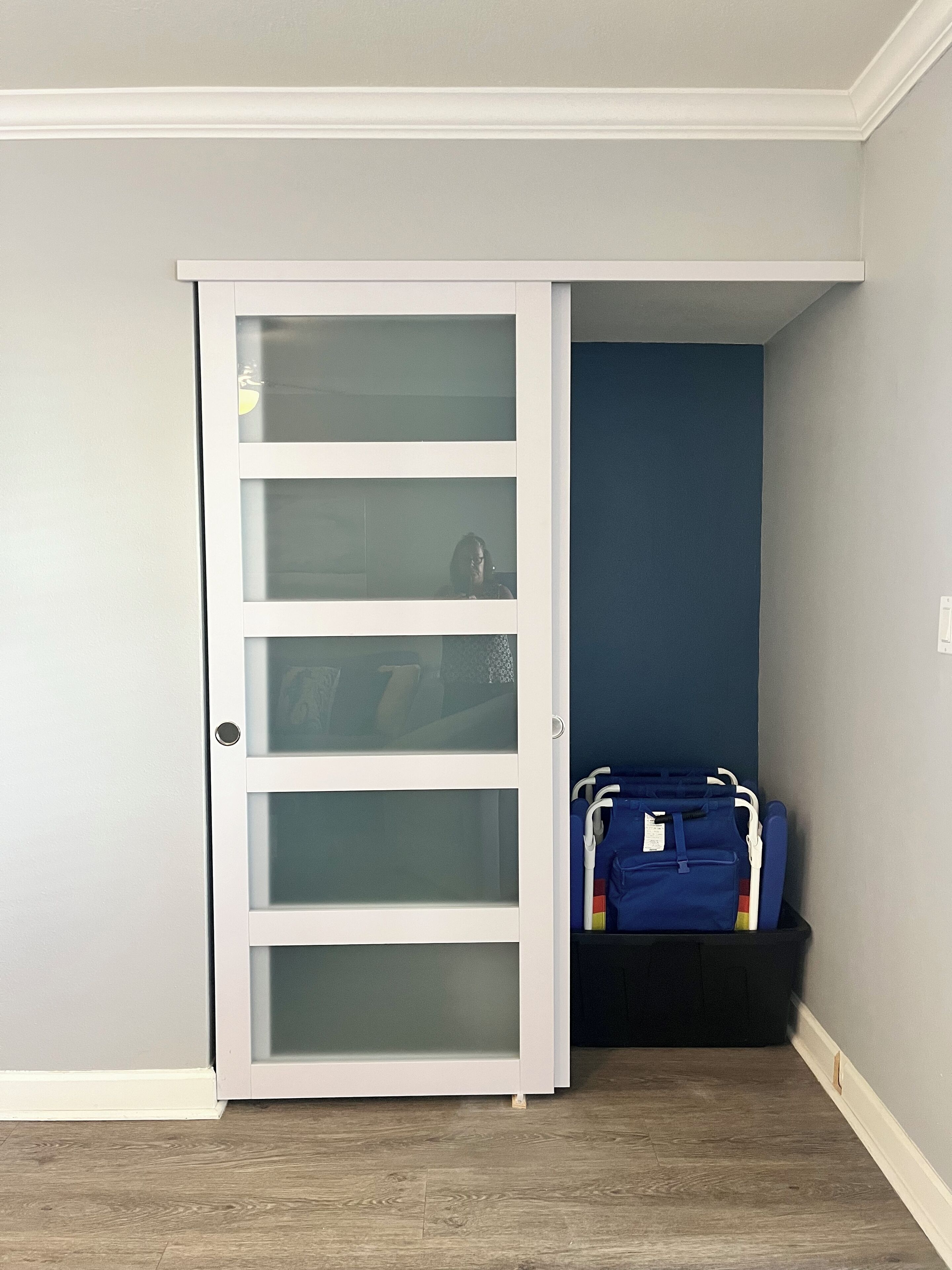 A room with a white door with frosted glass panels, a dark blue wall, and a black storage bin with blue bags.