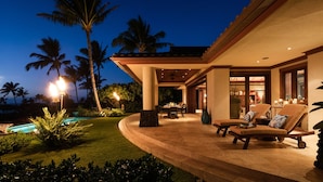 A well-lit outdoor patio area with a pool, lounge chairs, and a dining table. - Stunning Villa with Ocean Views, Pool, Home Theatre + Resort Amenities (Kamuela)