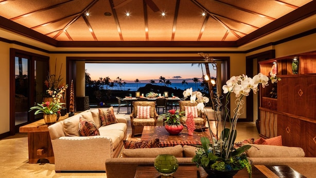 A spacious living room with a large window overlooking a scenic view, furnished with sofas, a coffee table, and decorative plants. - Stunning Villa with Ocean Views, Pool, Home Theatre + Resort Amenities (Kamuela)