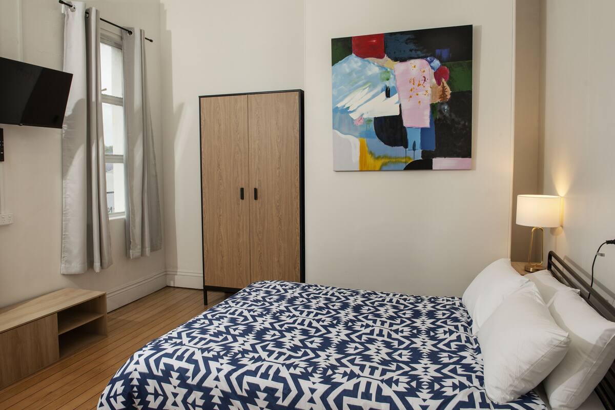 Quarto com cama, guarda-roupa, um quadro, televisão e abajur.