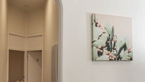 Un couloir doté d’une moquette sombre, d’un mur blanc orné d’un tableau représentant un cactus, et d’une porte donnant sur une pièce où se trouve un lit recouvert d’une couvre-lit à motifs.