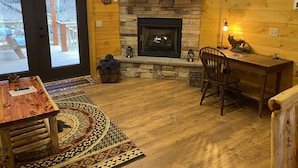 A cozy cabin interior with a stone fireplace, wooden walls, a TV mounted above it, and a dining area with a table and chairs. - New 2/2 Log Cabin, Rushing Trout Stream, Hot Tub, Fireplace, 8 Acres, WiFi (Deep Gap)