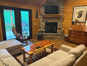 A cozy living room with a stone fireplace, a television mounted above it, a wooden coffee table, and a comfortable seating area with a sofa and a rocking chair.