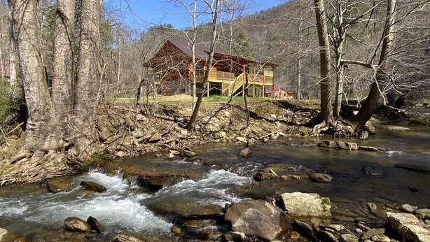A cabin with a wooden deck and stairs leading up to a porch, surrounded by a forest and a flowing stream. - New 2/2 Log Cabin, Rushing Trout Stream, Hot Tub, Fireplace, 8 Acres, WiFi (Deep Gap)