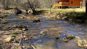 A cabin with a porch overlooking a clear stream, surrounded by trees. - New 2/2 Log Cabin, Rushing Trout Stream, Hot Tub, Fireplace, 8 Acres, WiFi (Deep Gap)