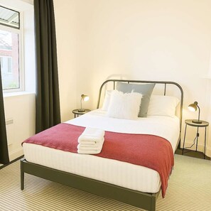 Een netjes opgemaakt bed met een rood-wit dekbedovertrek, twee nachtlampjes en een raam met gordijnen.