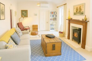 A cozy living room with a sofa, a wooden chest, a fireplace, a bookshelf, and a door leading to another room.