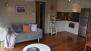 Um espaço de estar compacto com sofá, mesa redonda pequena, cozinha com armários e eletrodomésticos, e uma escada.