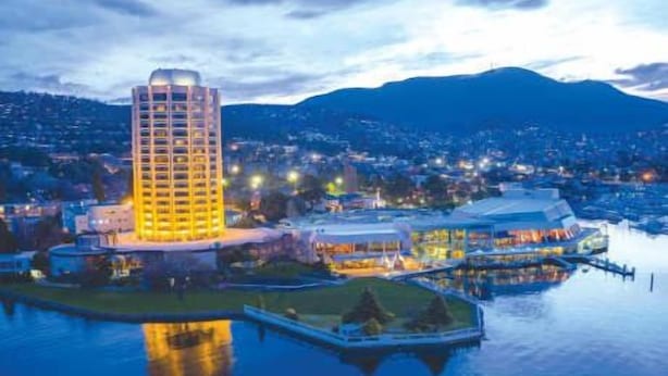 Um hotel moderno e imponente, localizado à beira de um corpo d'água, com vista para o horizonte urbano e montanhas ao fundo.