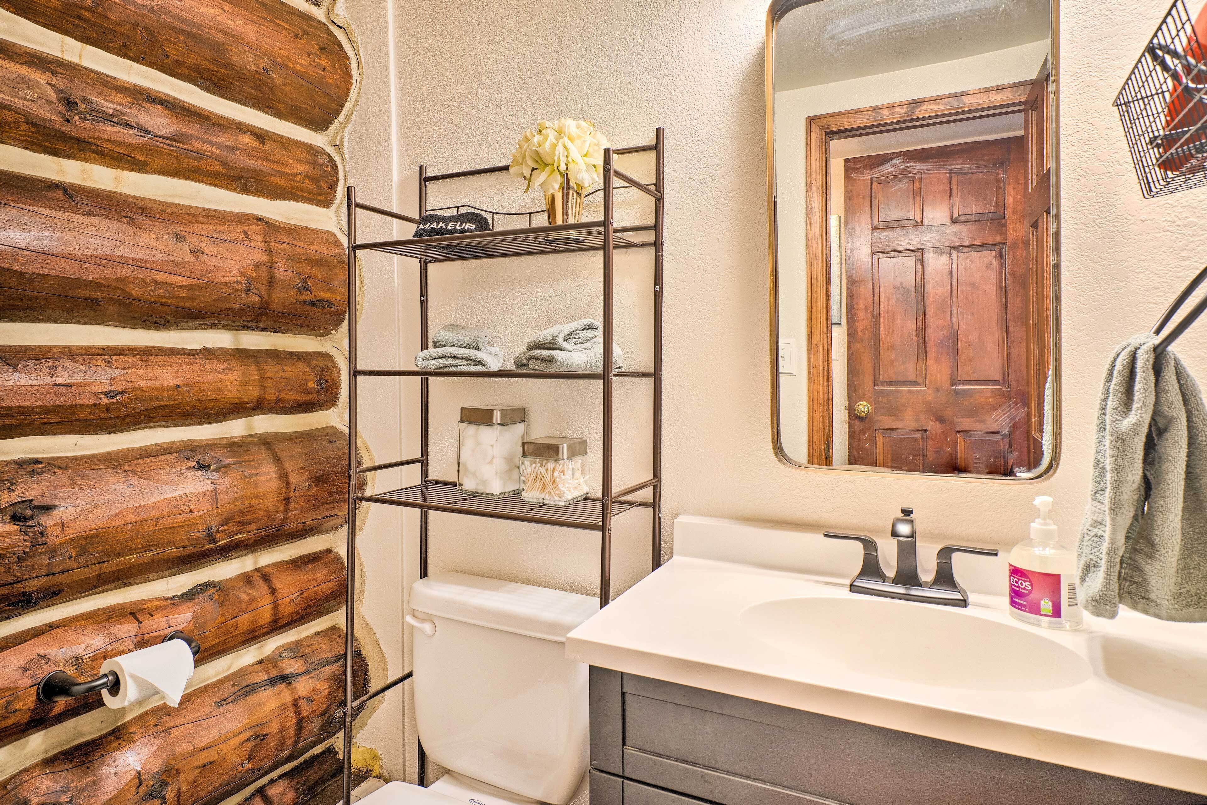 Un baño con pared de madera rústica, estante metálico con toallas y artículos de aseo, lavamanos con espejo y puerta de madera.