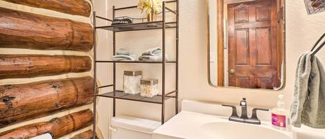 Un baño con pared de madera rústica, estante metálico con toallas y artículos de aseo, lavamanos con espejo y puerta de madera.