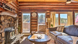 A cozy cabin interior with a stone fireplace, a vintage television, a leather sofa, and a large window offering a scenic view. - Classic Colorado Log Home w/ Mountain Views! (Florissant)