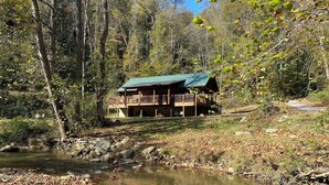Une cabane dotée d’une terrasse et d’un porche abrité, entourée d’arbres et bordée par un ruisseau.