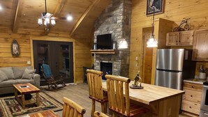 L’intérieur chaleureux d’un chalet avec une table à manger en bois, des chaises, une cheminée, un coin cuisine équipé d’un réfrigérateur et un salon avec un canapé et un fauteuil à bascule.