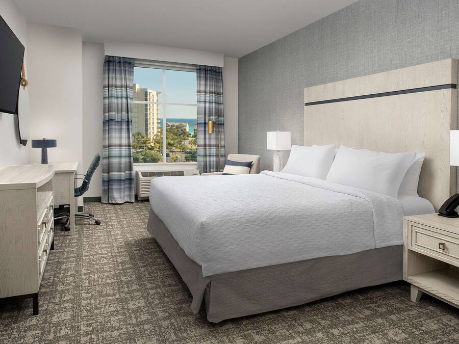 A hotel room with a large bed, a desk, a chair, a TV, and a view of the city.