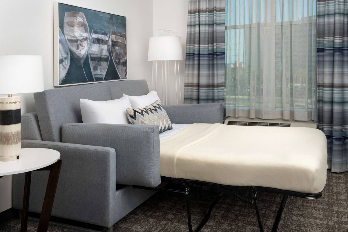 Ein modernes Hotelzimmer mit grauem Sofa, einem kleinen weißen Tisch, einem Nachttisch, einem weißen Bett, einem großen Fenster mit Vorhängen und einem Bild an der Wand.