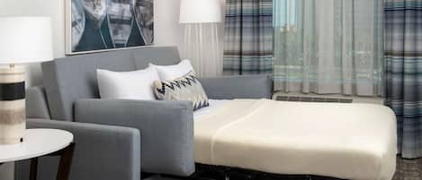 A modern hotel room with a grey sofa, a small white table, a bedside table, a white bed, a large window with curtains, and a painting on the wall.