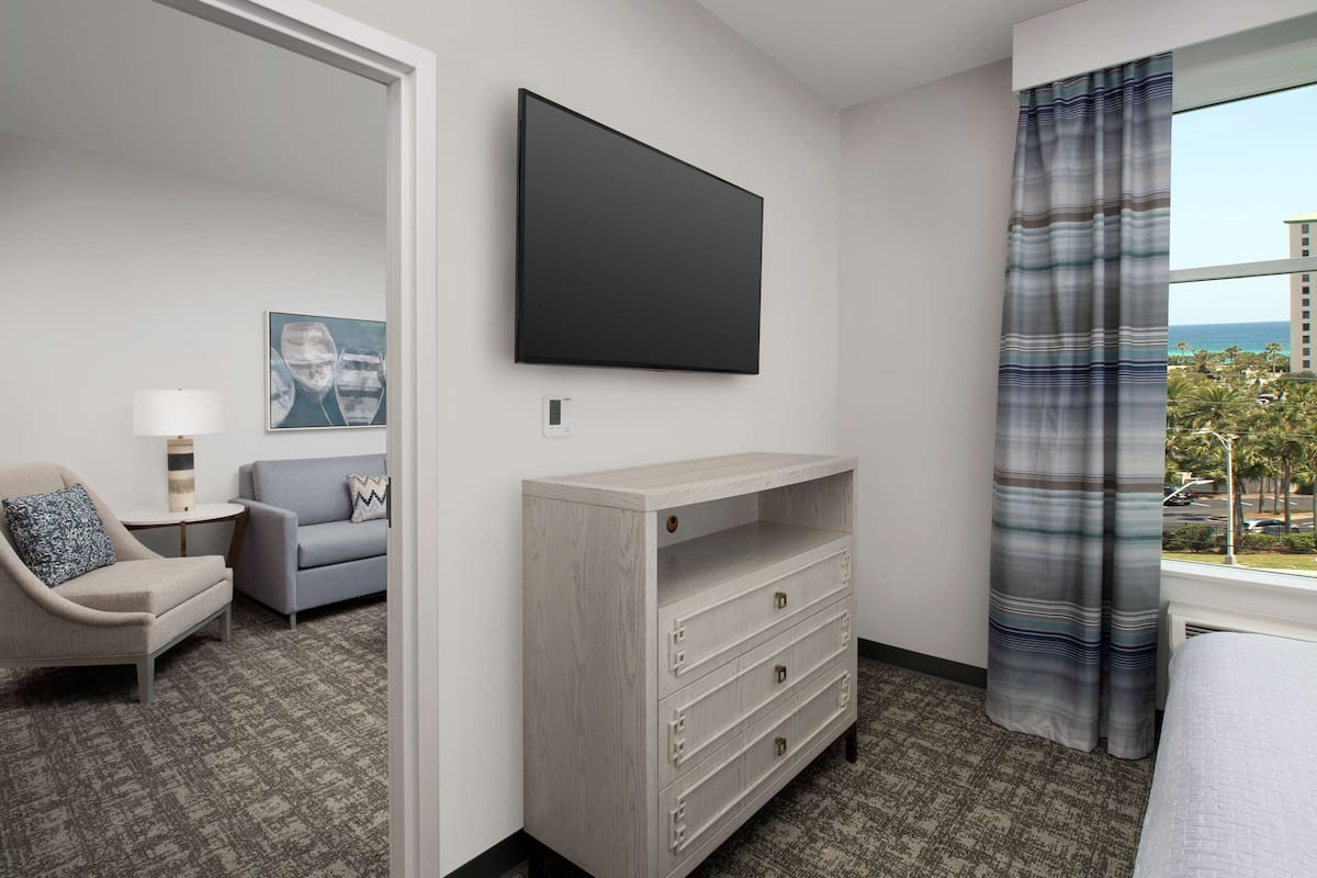 Ein Hotelzimmer mit Flachbildfernseher an der Wand, einer grauen Kommode mit Schubladen, einem grauen Sofa mit einem gemusterten Kissen, einem kleinen Beistelltisch mit Lampe und einem Fenster mit Blick auf Palmen und Gebäude.
