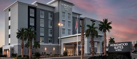A modern hotel with a prominent entrance, palm trees, and a sign indicating it is a Homewood Suites by Hilton.
