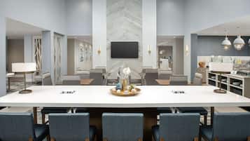 A modern hotel conference room with a large rectangular table, six upholstered chairs, a flat-screen TV mounted on the wall, and a kitchen area with a sink and countertop.