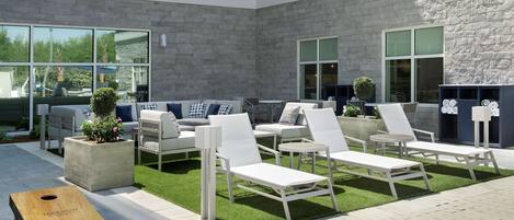 A modern outdoor lounge area with white lounge chairs, a concrete planter with a small tree, and a wooden board game table.