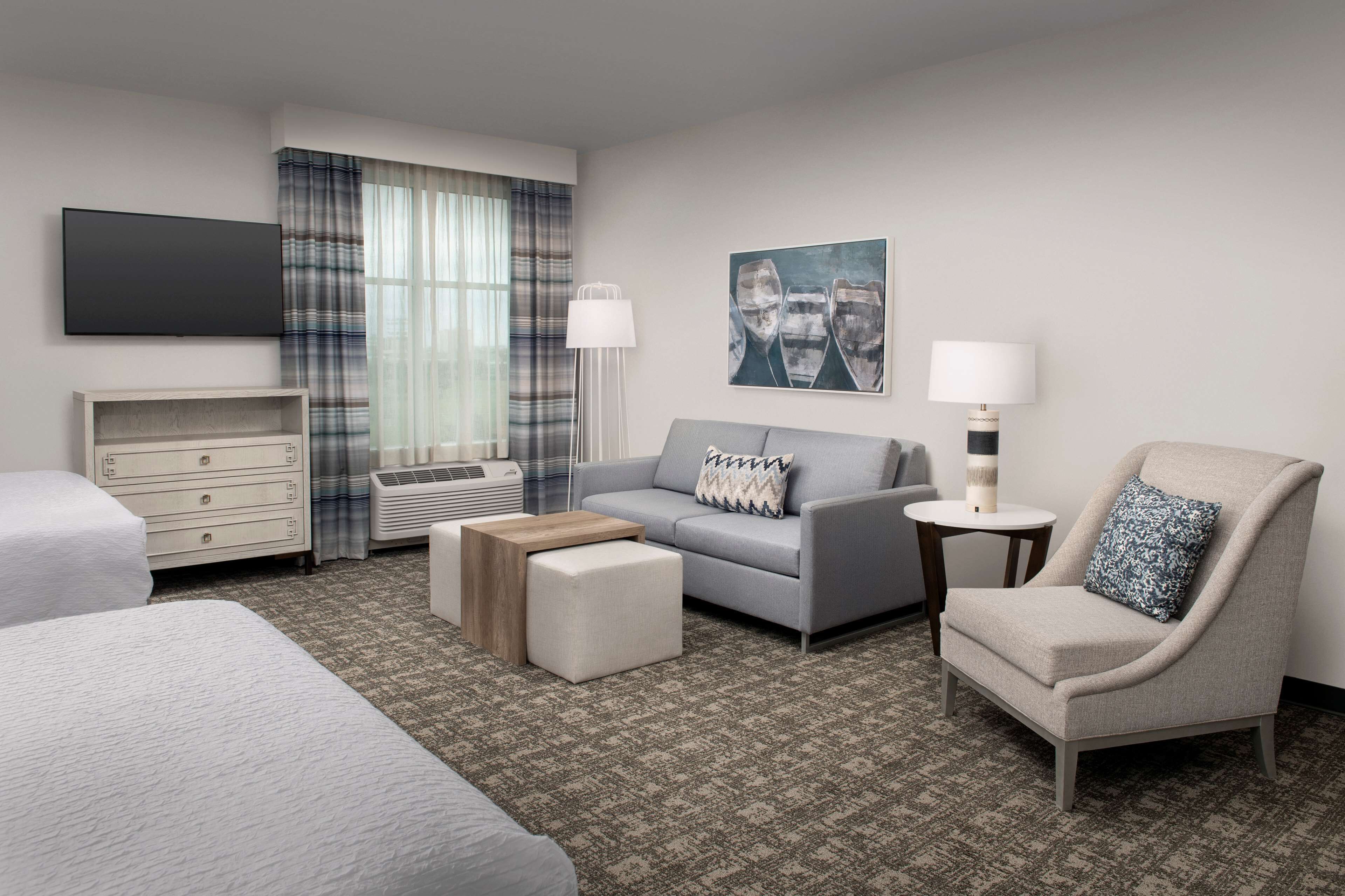 A hotel room with a bed, a sofa, a chair, a small table, a lamp, and a painting on the wall.