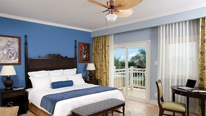Ein Hotelzimmer mit einem großen Bett, einem Deckenventilator, einem Balkon mit weißer Reling und einem Schreibtisch mit Stuhl.