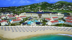 Un resort con diversi edifici alberghieri, una vasta area piscina e una spiaggia con numerosi ombrelloni.