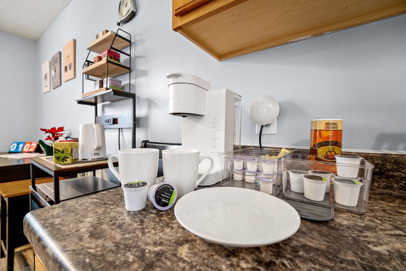 Une cuisine équipée d’une machine à café, de tasses, d’une assiette et de récipients contenant du café en poudre ou du liquide pour café.