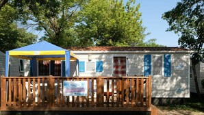 Una casa móvil con terraza amurallada, un toldo azul y amarillo, y un letrero que dice 'Happy Camp'.